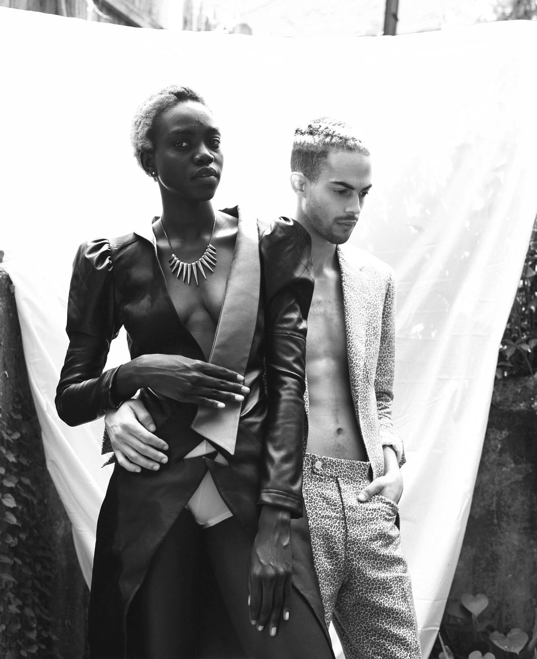 Visionarios Magazine editorial: the pair side by side, the leather coat and the leopard suit worn open, in an overgrown courtyard